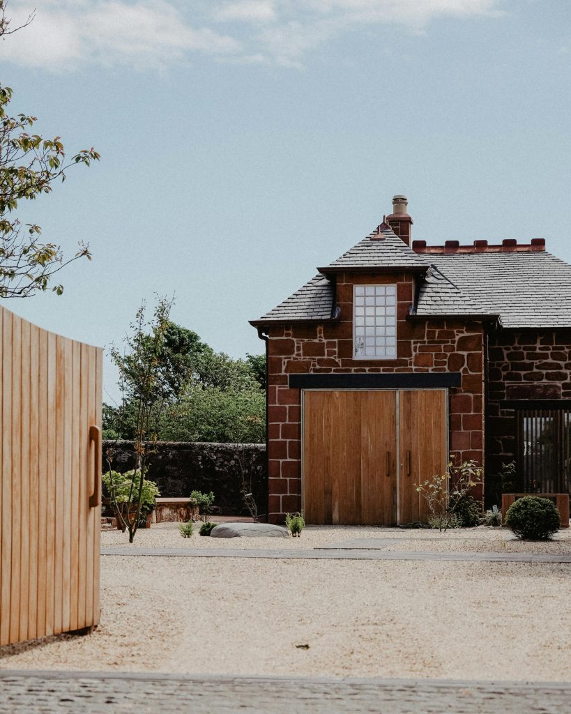 Rustic brick house with wooden doors, surrounded by a gravel yard and greenery under a clear blue sky.