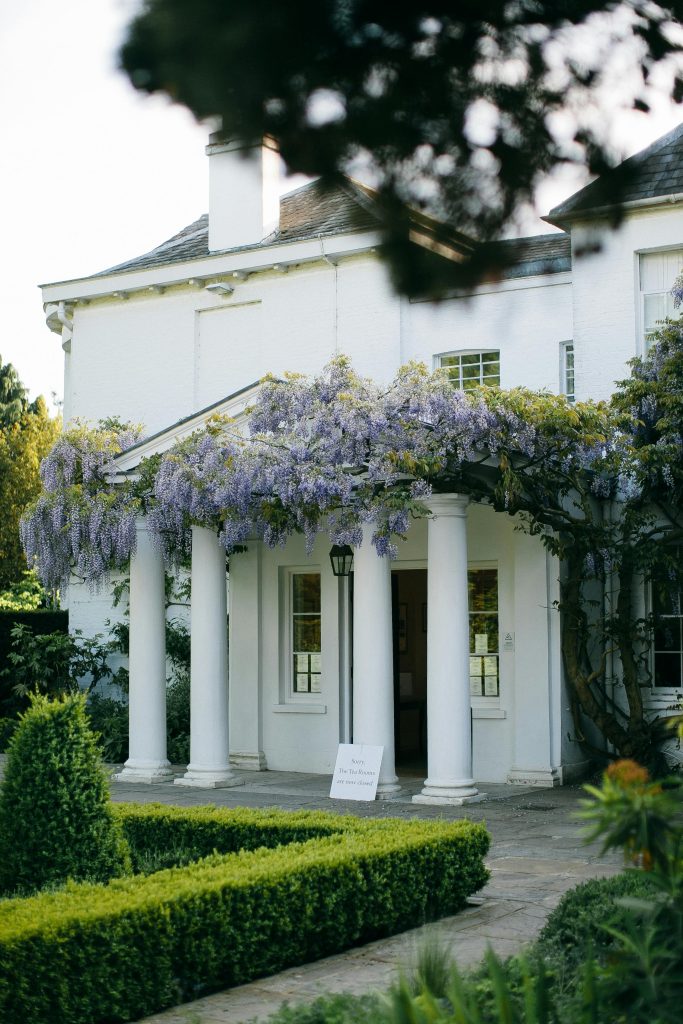 Elegant white house with wisteria-draped columns, surrounded by lush greenery.