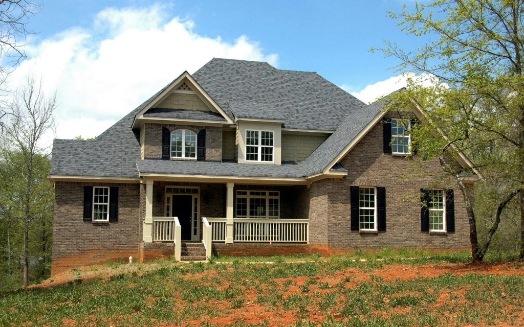 Two-story brick house with large windows, front porch, and gabled roof set against a clear blue sky and lush greenery.